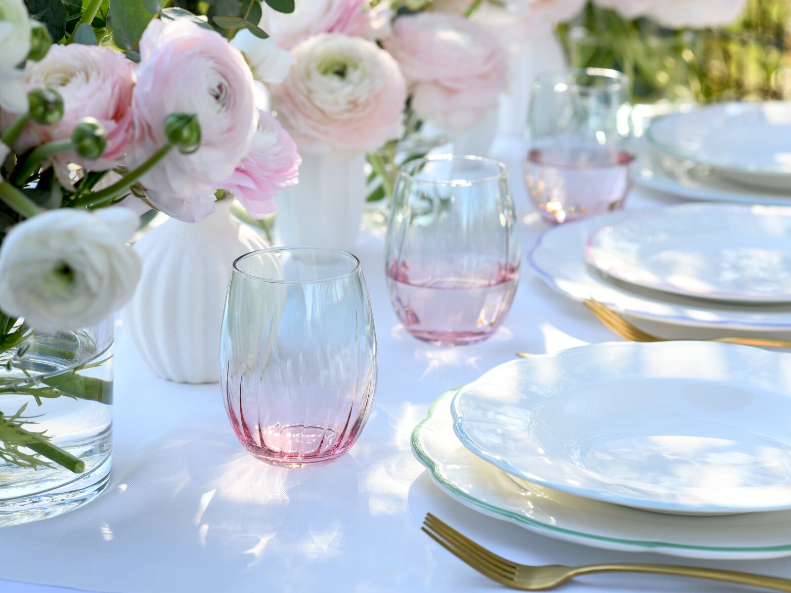Eleganter gedeckter Tisch mit roséfarbenen Trinkgläsern, weißen Tellern mit gewelltem Rand, goldfarbenem Besteck und rosa-weißen Blumen in Vasen, aufgenommen in leicht schräger Seitenansicht auf Tischhöhe.