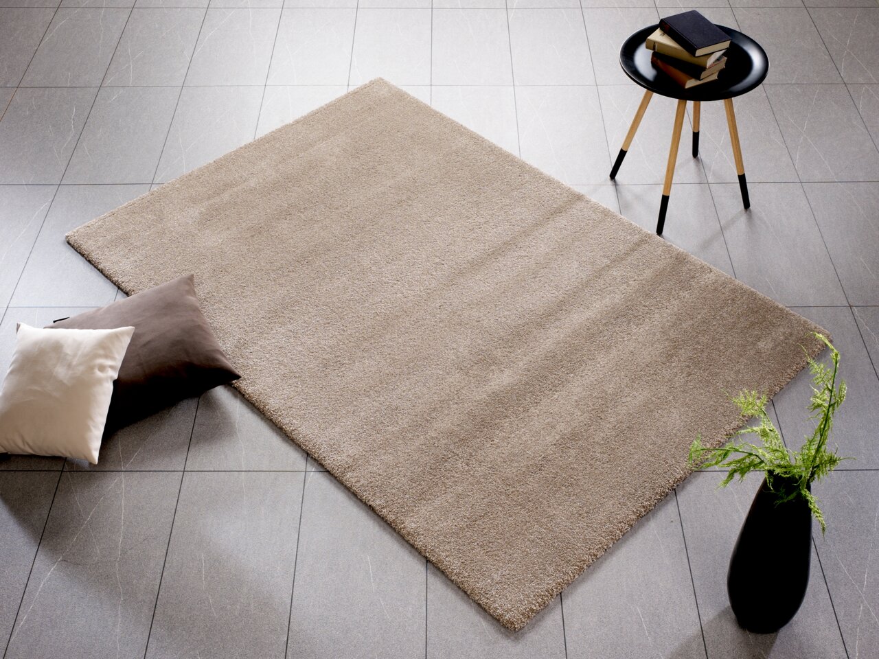 Beige Uni-Teppich auf grauem Fliesenboden, umgeben von zwei Kissen, einem Beistelltisch mit Büchern und einer Vase mit Pflanzen, aus der Vogelperspektive fotografiert.