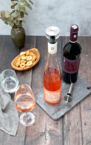 Ausgießer und Verschluss für Champagnerflaschen, von oben fotografiert, neben einer Flasche Roséwein und einer Flasche Rotwein auf einem Tisch mit Snacks und Gläsern.