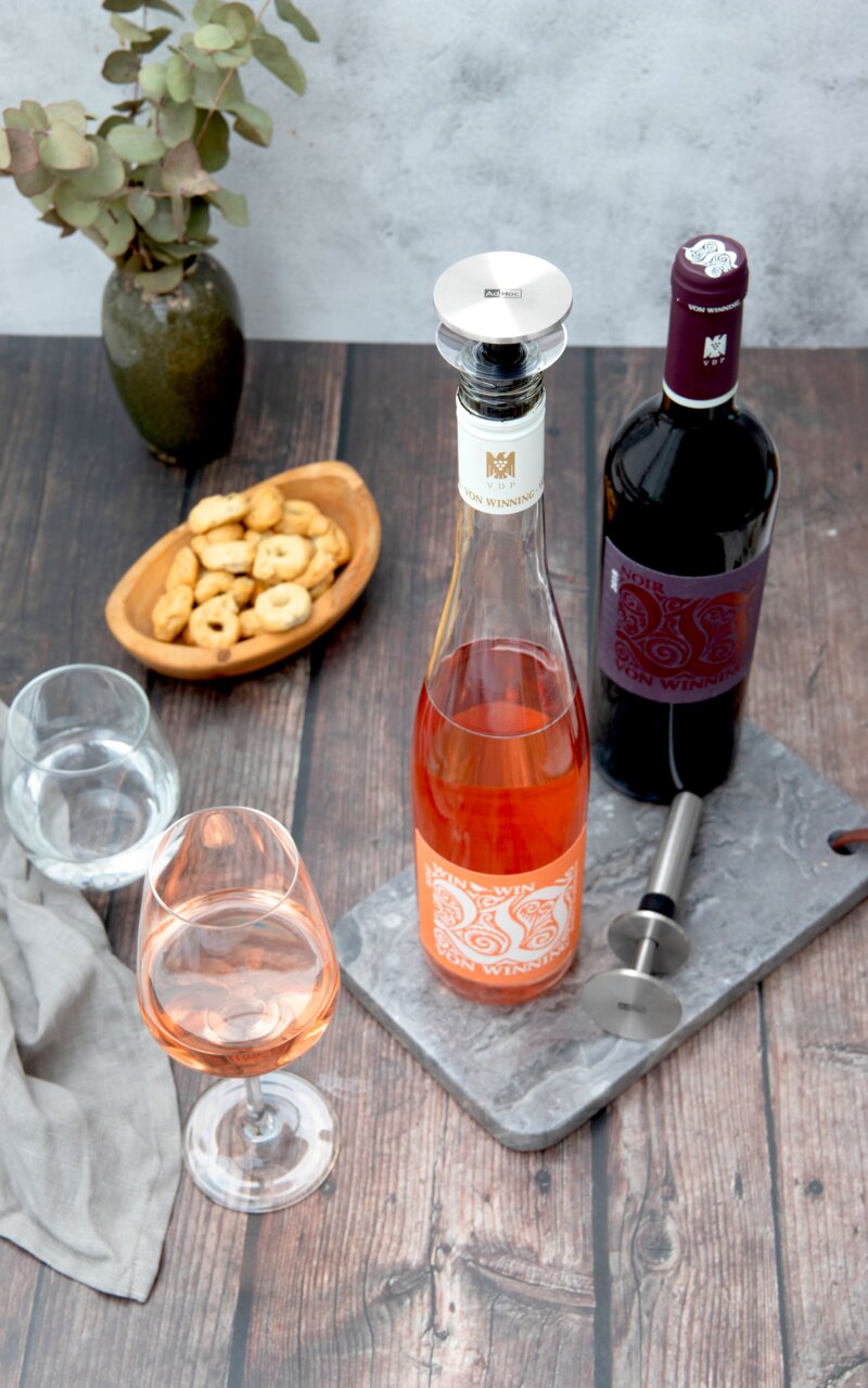Een schenktuit en een dop voor champagneflessen, gefotografeerd van bovenaf, naast een fles rosé en een fles rode wijn op een tafel met hapjes en glazen.