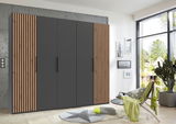 Moderner 5-türiger Drehtürenschrank in dunklem Grau mit Holzlamellen an den Seiten, frontal fotografiert in einem hellen Raum mit großen Fenstern.
