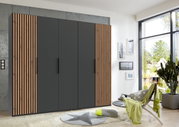 Moderner 5-türiger Drehtürenschrank in dunklem Grau mit Holzlamellen an den Seiten, frontal fotografiert in einem hellen Raum mit großen Fenstern.