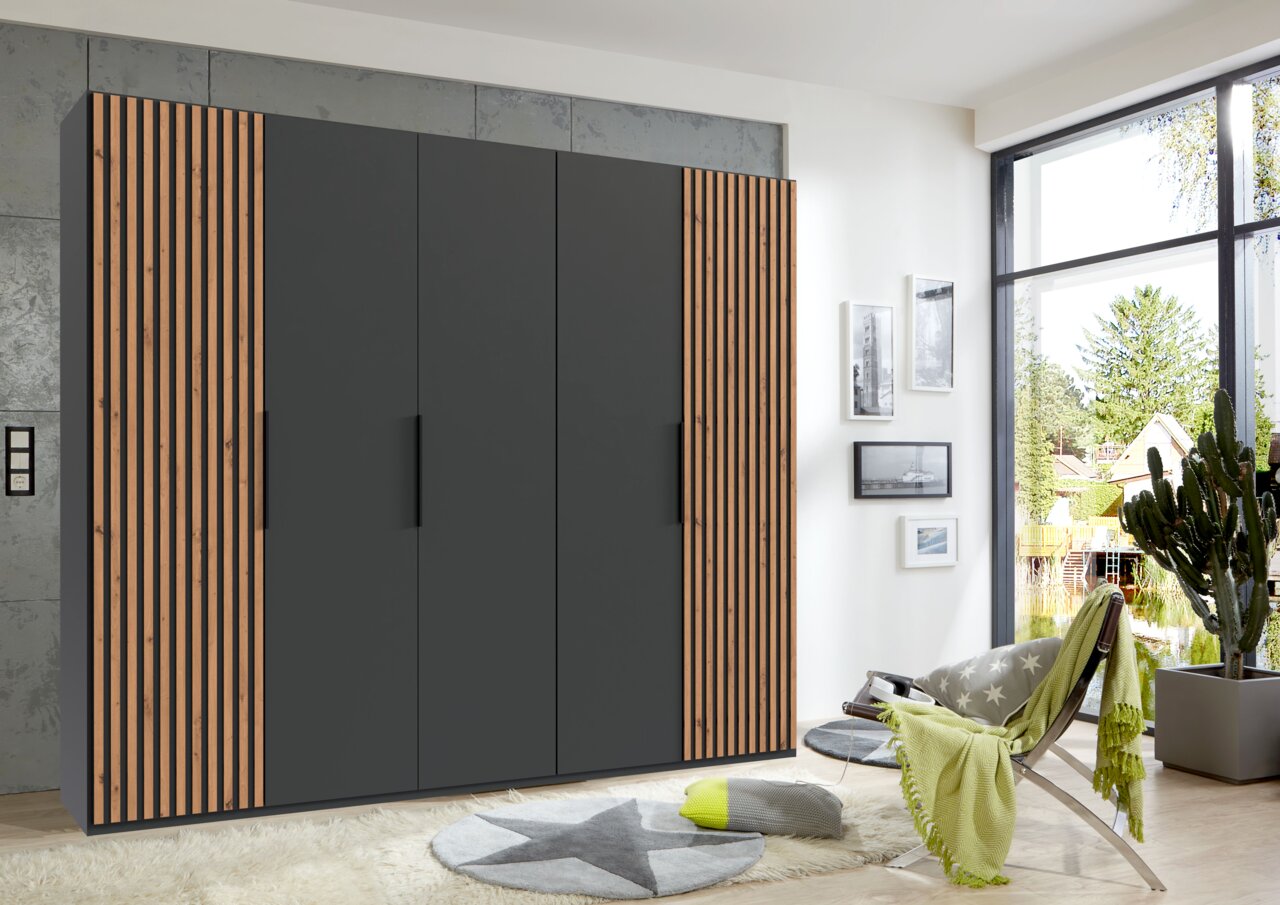Z2 Drehtürenschrank 5-türig ANDORRA Moderner 5-türiger Drehtürenschrank in dunklem Grau mit Holzlamellen an den Seiten, frontal fotografiert in einem hellen Raum mit großen Fenstern.