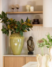 Ein stilvolles Arrangement mit einem großen, grünen Keramiktopf mit Pflanzen, einer modernen Skulptur und Teelichtern auf einem Regal. Perspektive: Frontalansicht.