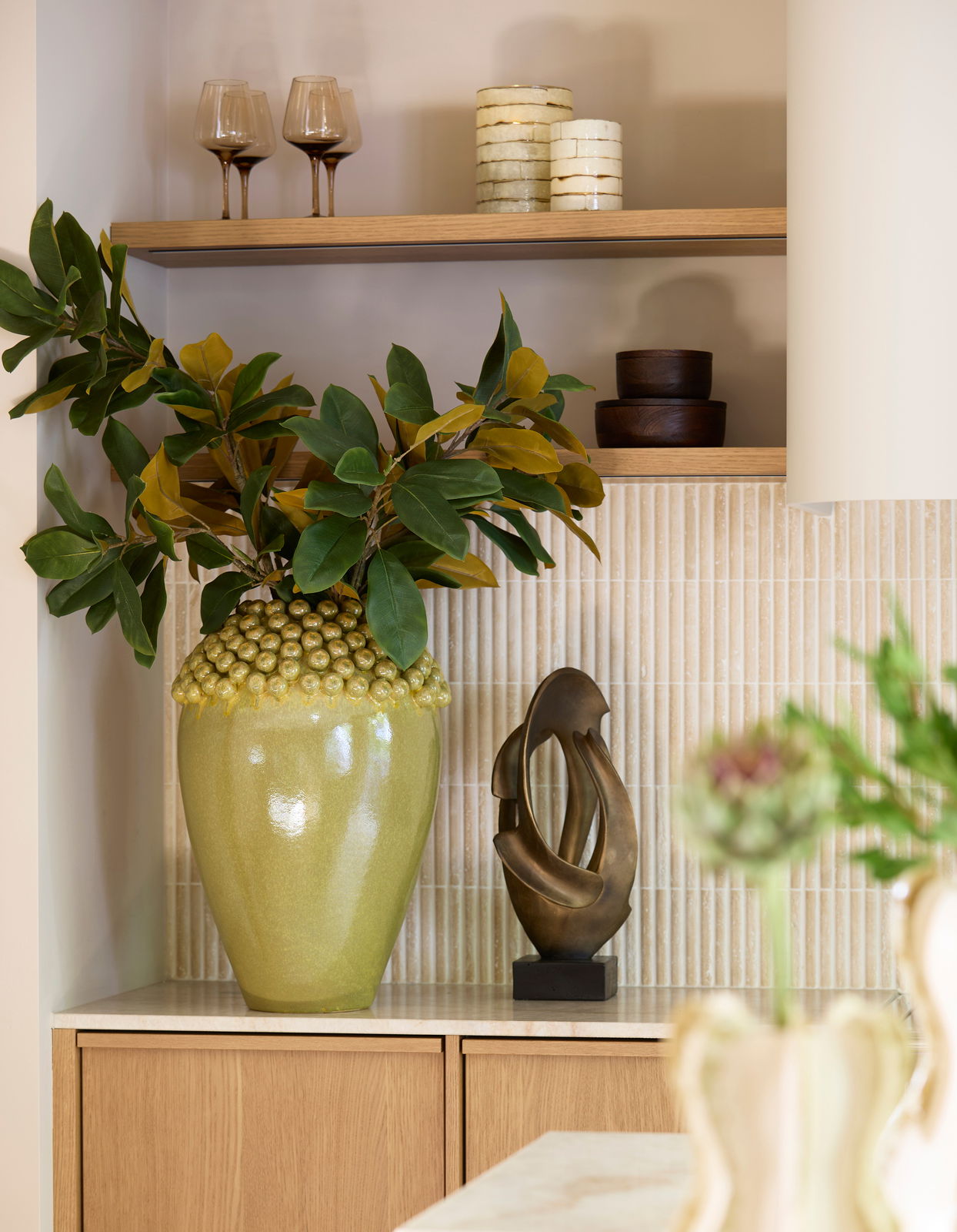 Ein stilvolles Arrangement mit einem großen, grünen Keramiktopf mit Pflanzen, einer modernen Skulptur und Teelichtern auf einem Regal. Perspektive: Frontalansicht.