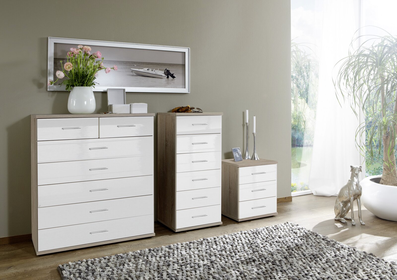 XORA Schubkastenkommode MALTA Drei moderne Schubkastenkommoden in verschiedenen Größen mit weiß glänzenden Fronten und Holzrahmen, dekoriert mit Blumen und Kerzen, in einem hellen Raum mit Teppich und Pflanzen, seitliche Perspektive.