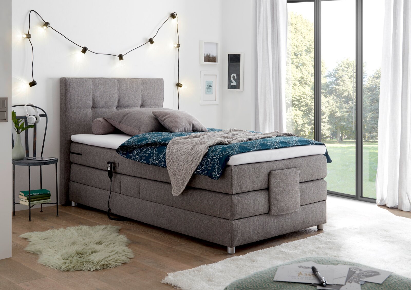 Esposa Boxspringbett MANOLO Graues Boxspringbett in einem modernen Schlafzimmer, seitliche Perspektive, mit dekorativen Kissen und Decke, umgeben von Lichterkette und großen Fenstern.