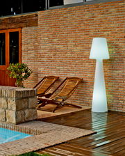 LED Outdoor Stehlampe Lola 165, beleuchtet, auf einer Terrasse neben zwei Holzliegen und einer Ziegelwand, seitliche Perspektive