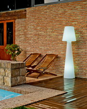 LED Outdoor Stehlampe Lola 165, beleuchtet, auf einer Terrasse neben zwei Holzliegen und einer Ziegelwand, seitliche Perspektive