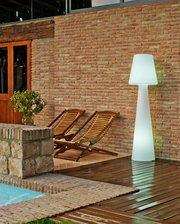 LED Outdoor Stehlampe Lola 165, beleuchtet, auf einer Terrasse neben zwei Holzliegen und einer Ziegelwand, seitliche Perspektive