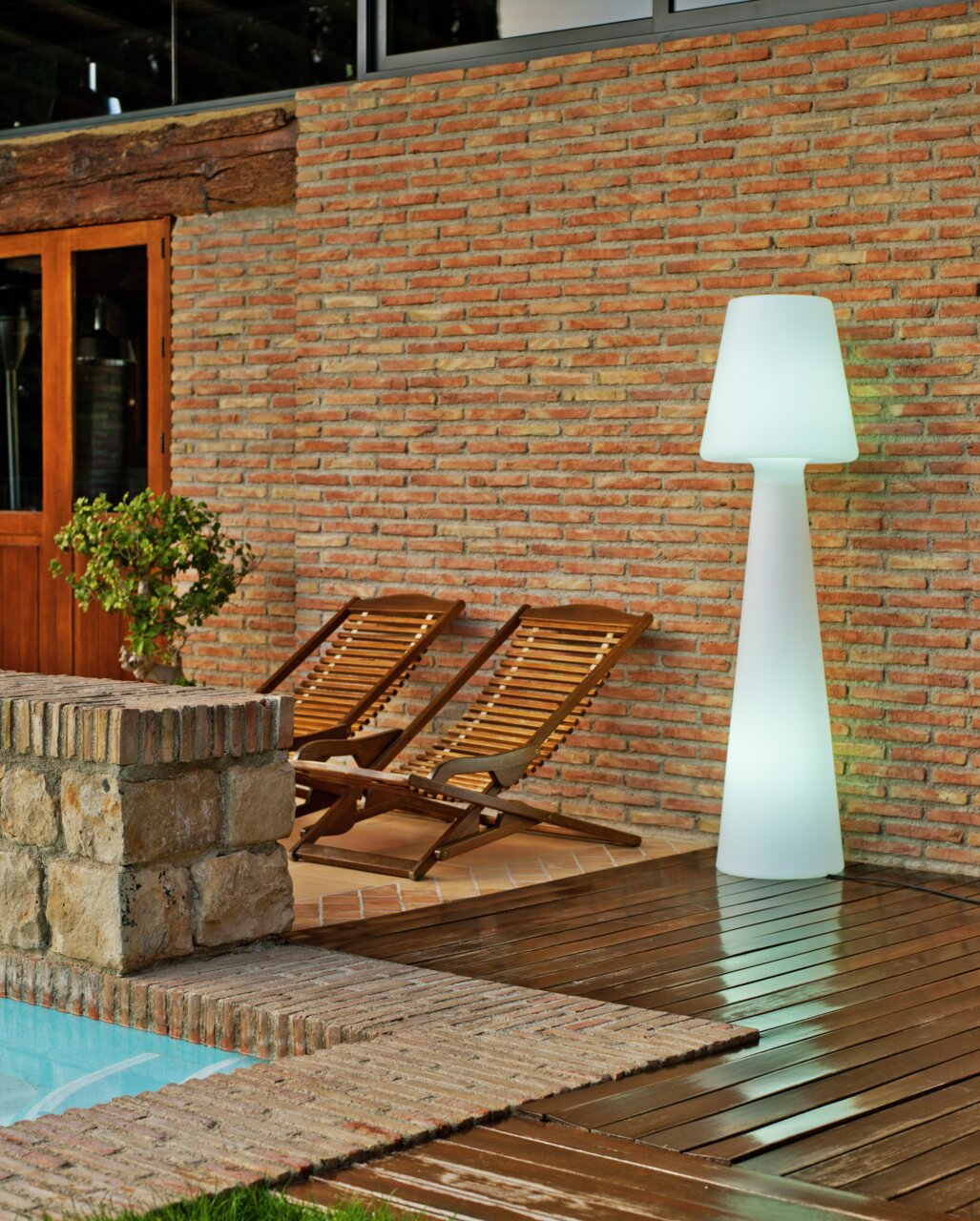 LED-buitenstaande lamp Lola 165, verlicht, op een terras naast twee houten ligstoelen en een bakstenen muur, zijaanzicht