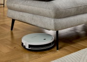 Ronde stofzuigrobot in wit en zwart maakt de parketvloer schoon onder een grijze bank; zijaanzicht van dicht bij de vloer vanuit een licht schuin perspectief.