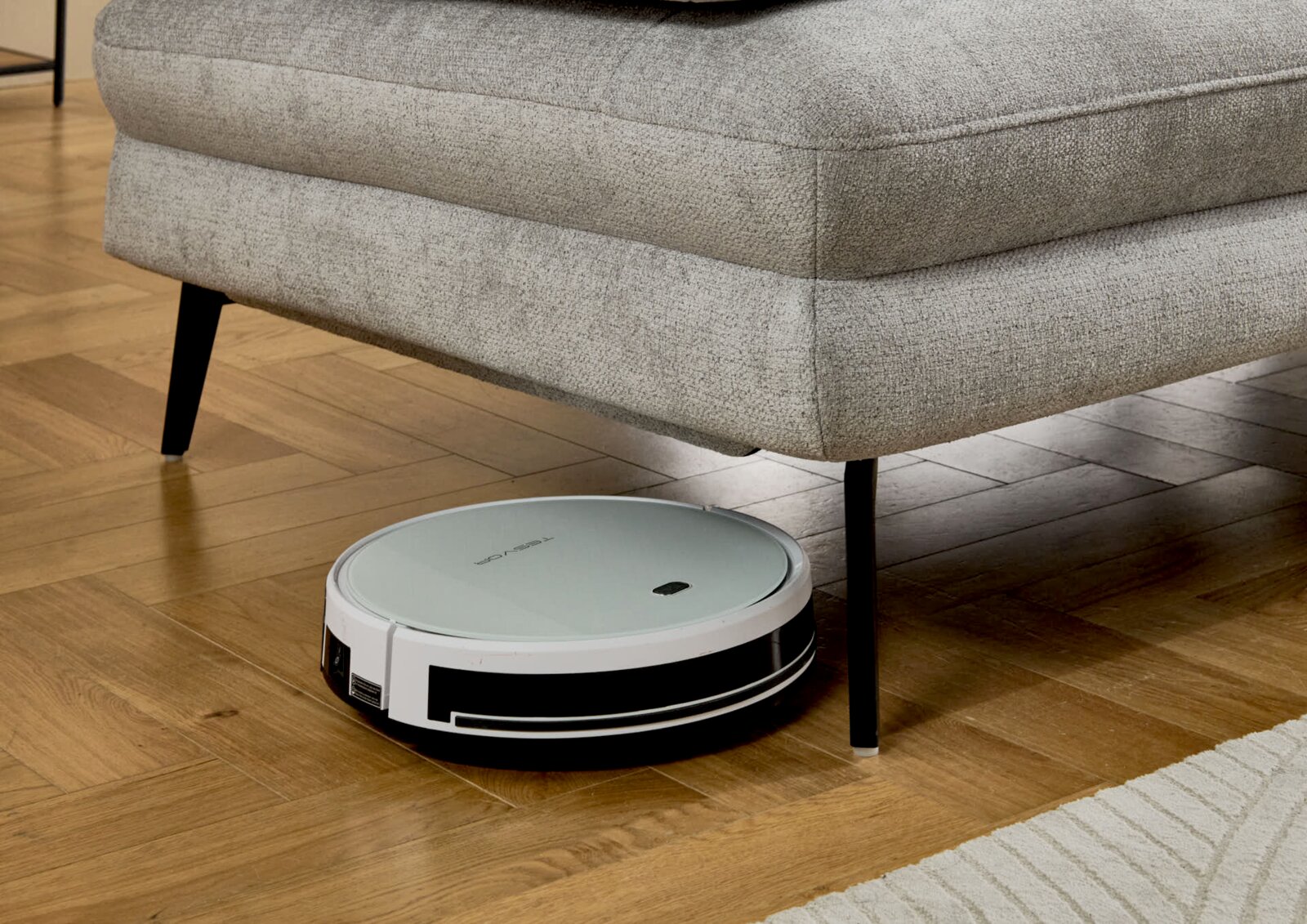 Ronde stofzuigrobot in wit en zwart maakt de parketvloer schoon onder een grijze bank; zijaanzicht van dicht bij de vloer vanuit een licht schuin perspectief.