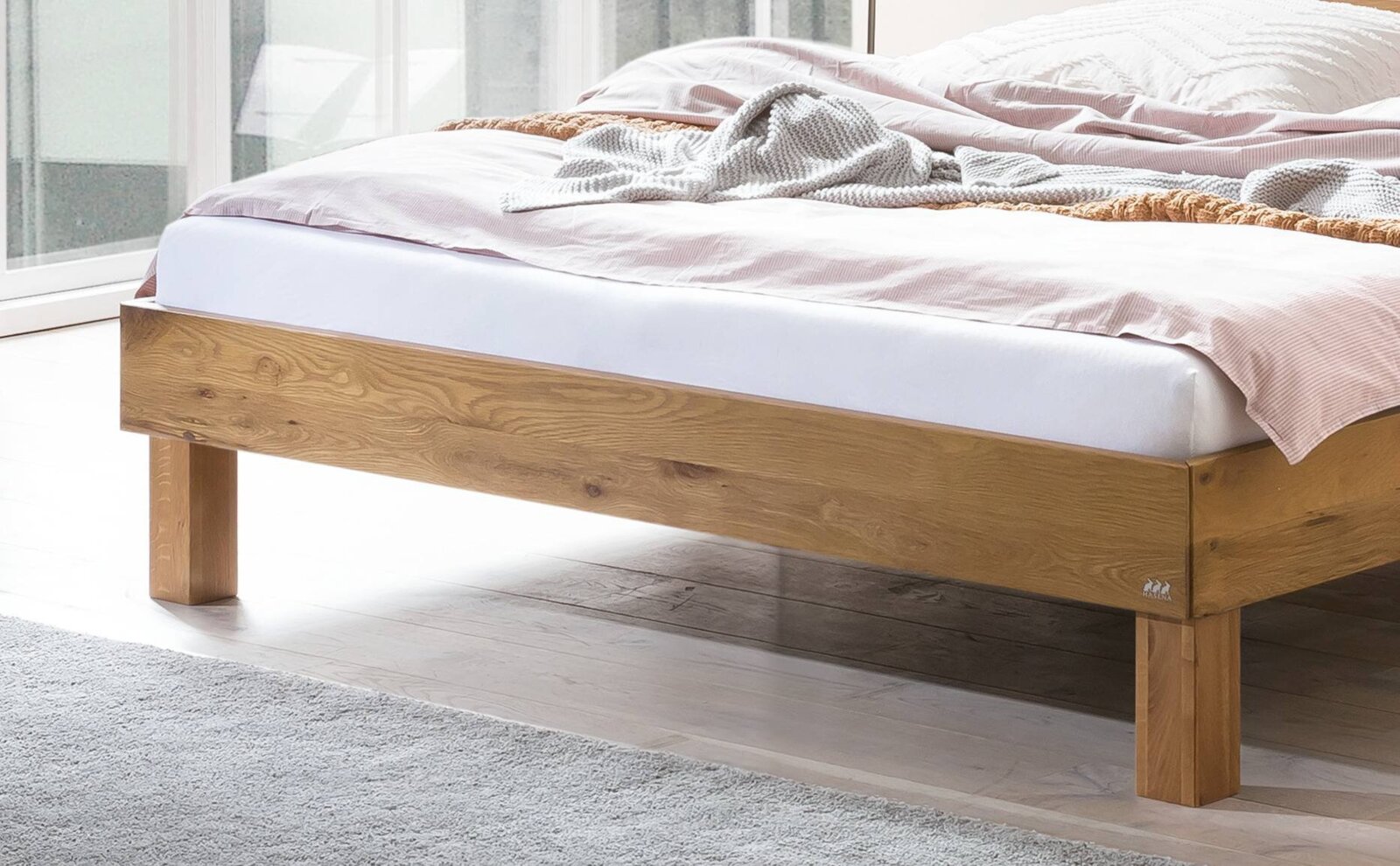 Massives Holzbettgestell in Eiche-Optik mit sichtbarer Maserung und stabilen eckigen Beinen, gezeigt in seitlicher Perspektive im hellen Schlafzimmer.