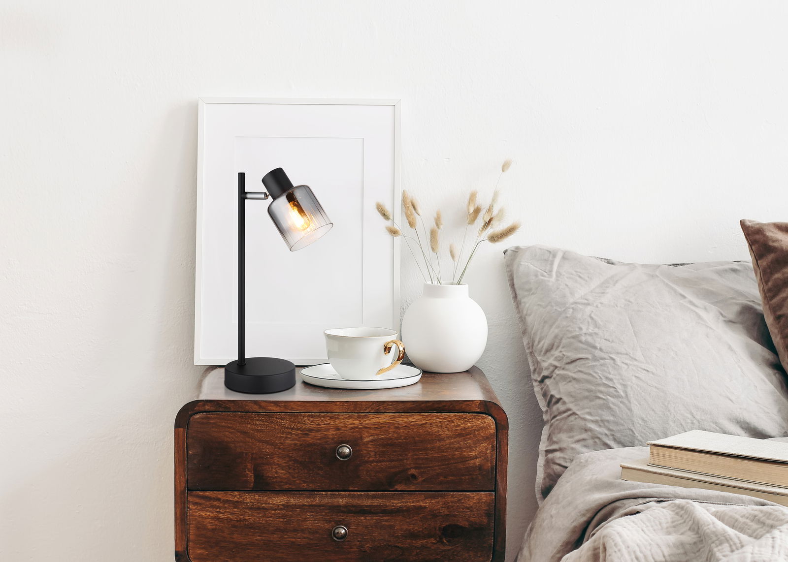 NOWA Tischleuchte JACK Moderne Tischleuchte mit schwarzem Gestell und rauchigem Glasschirm auf einem Nachttisch, seitliche Perspektive.