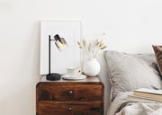 Moderne Tischleuchte mit schwarzem Gestell und rauchigem Glasschirm auf einem Nachttisch, seitliche Perspektive.