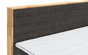 Nahaufnahme eines Boxspringbettes mit grauem Stoffkopfteil und Holzrahmen, seitliche Perspektive