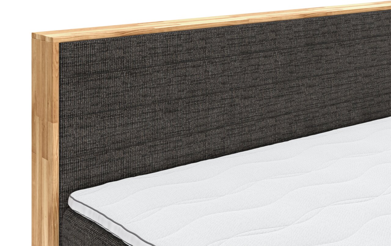 Nahaufnahme eines Boxspringbettes mit grauem Stoffkopfteil und Holzrahmen, seitliche Perspektive