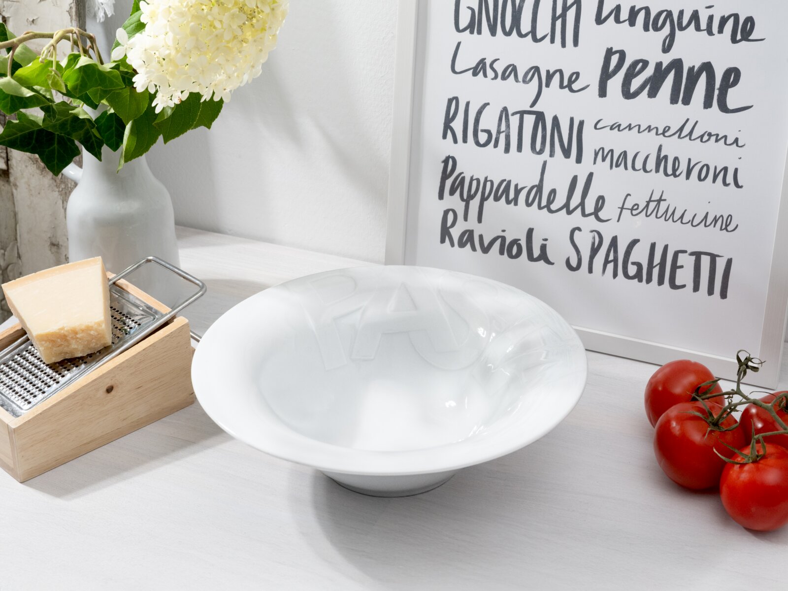 Weiße, tiefe Pastaschale mit eingeprägtem Schriftzug, schräg von vorne auf einem hellen Tisch fotografiert; daneben Käsereibe mit Parmesan, rote Tomaten und ein gerahmtes Pasta-Poster im Hintergrund.