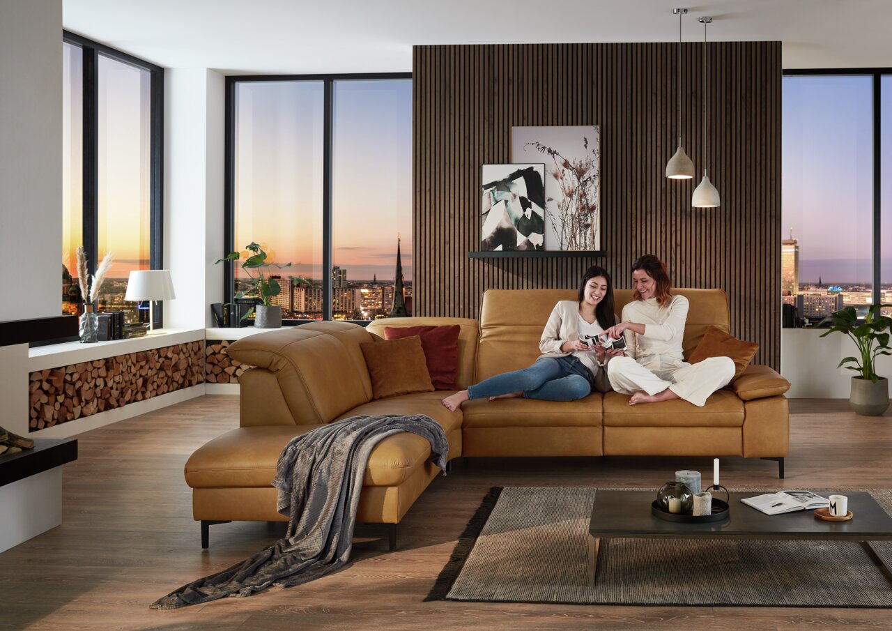 Gemütliche Polsterrundecke in einem modernen Wohnzimmer mit zwei Personen, die auf dem Sofa sitzen und ein Buch lesen. Die Perspektive zeigt das Sofa von vorne mit einem großen Fenster im Hintergrund, das einen Blick auf die Stadt bei Sonnenuntergang biet