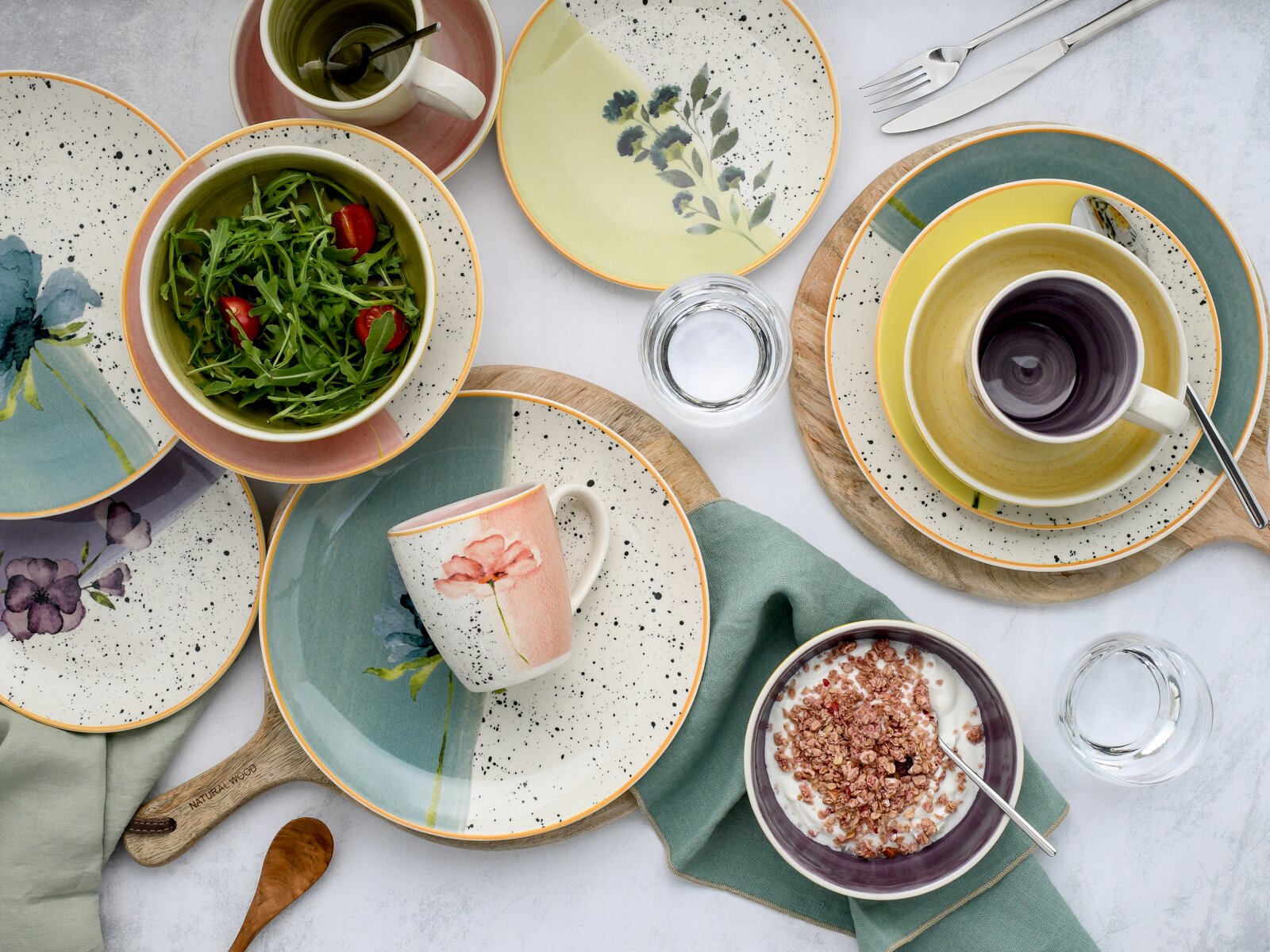 Buntes Keramikgeschirr mit floralen Motiven, Sprenkeldekor und orangefarbenen Rändern, als gedeckter Frühstückstisch mit Salat, Müsli und Getränken aus der Vogelperspektive arrangiert