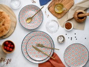 Draufsicht auf ein stilvolles Tisch-Arrangement mit gemustertem Keramikgeschirr (Teller und Schale in Weiß mit blau-rotem Sternmuster), goldfarbenem Besteck, Brot, Kirschtomaten, Gewürzen und Öl auf Holzbrett.