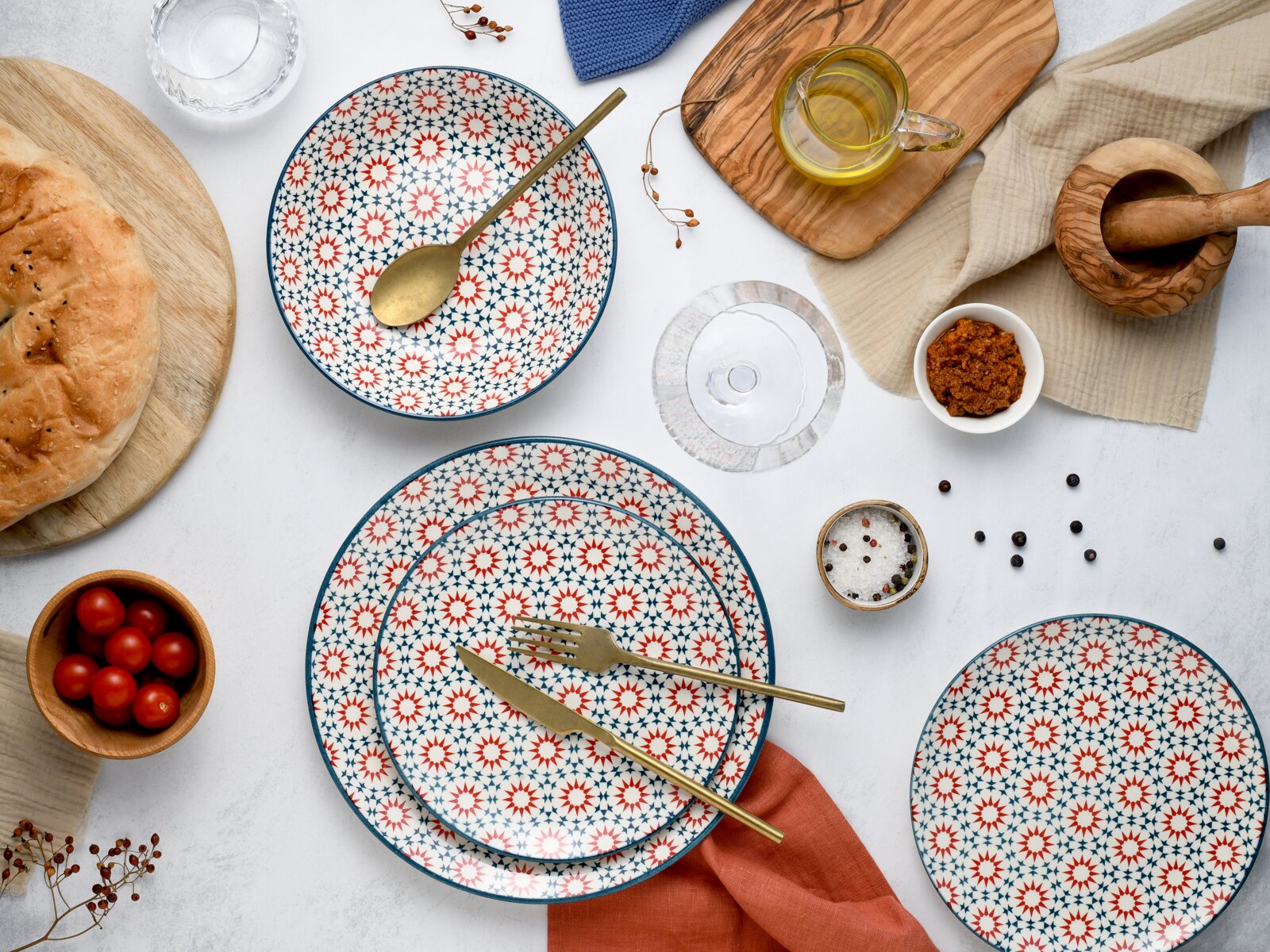 Draufsicht auf ein stilvolles Tisch-Arrangement mit gemustertem Keramikgeschirr (Teller und Schale in Weiß mit blau-rotem Sternmuster), goldfarbenem Besteck, Brot, Kirschtomaten, Gewürzen und Öl auf Holzbrett.