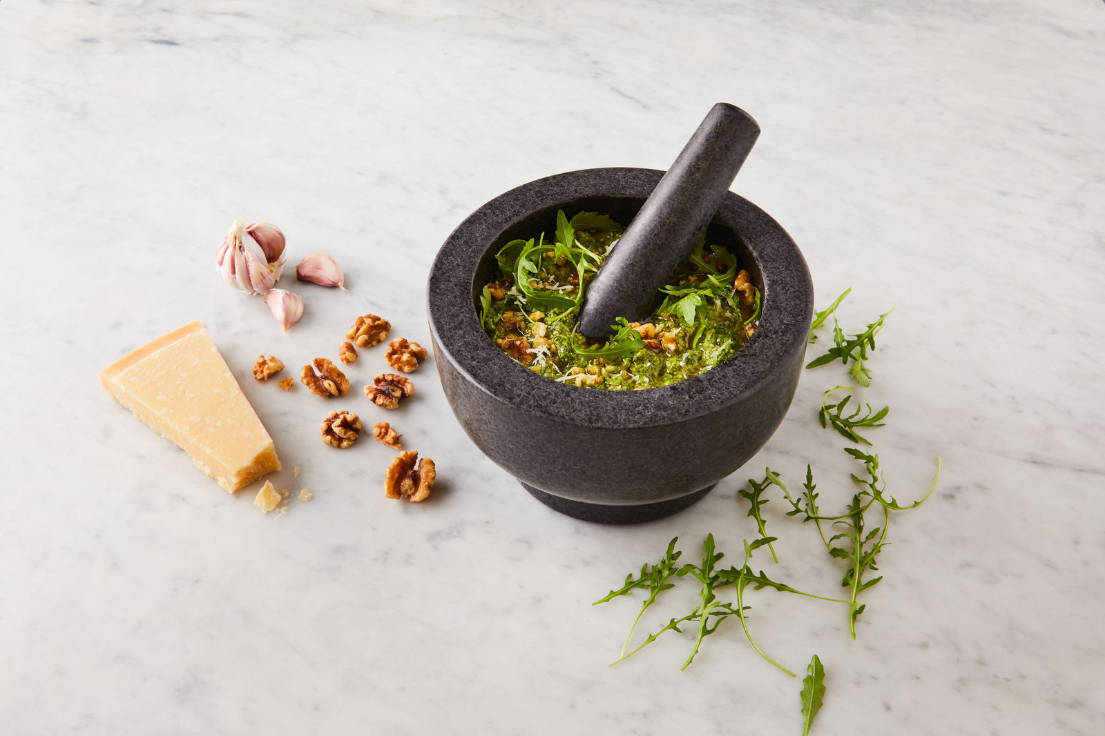 Mörser und Stößel aus dunklem Stein, gefüllt mit frischen Zutaten wie Rucola und Walnüssen, auf einer Marmoroberfläche. Perspektive von oben.