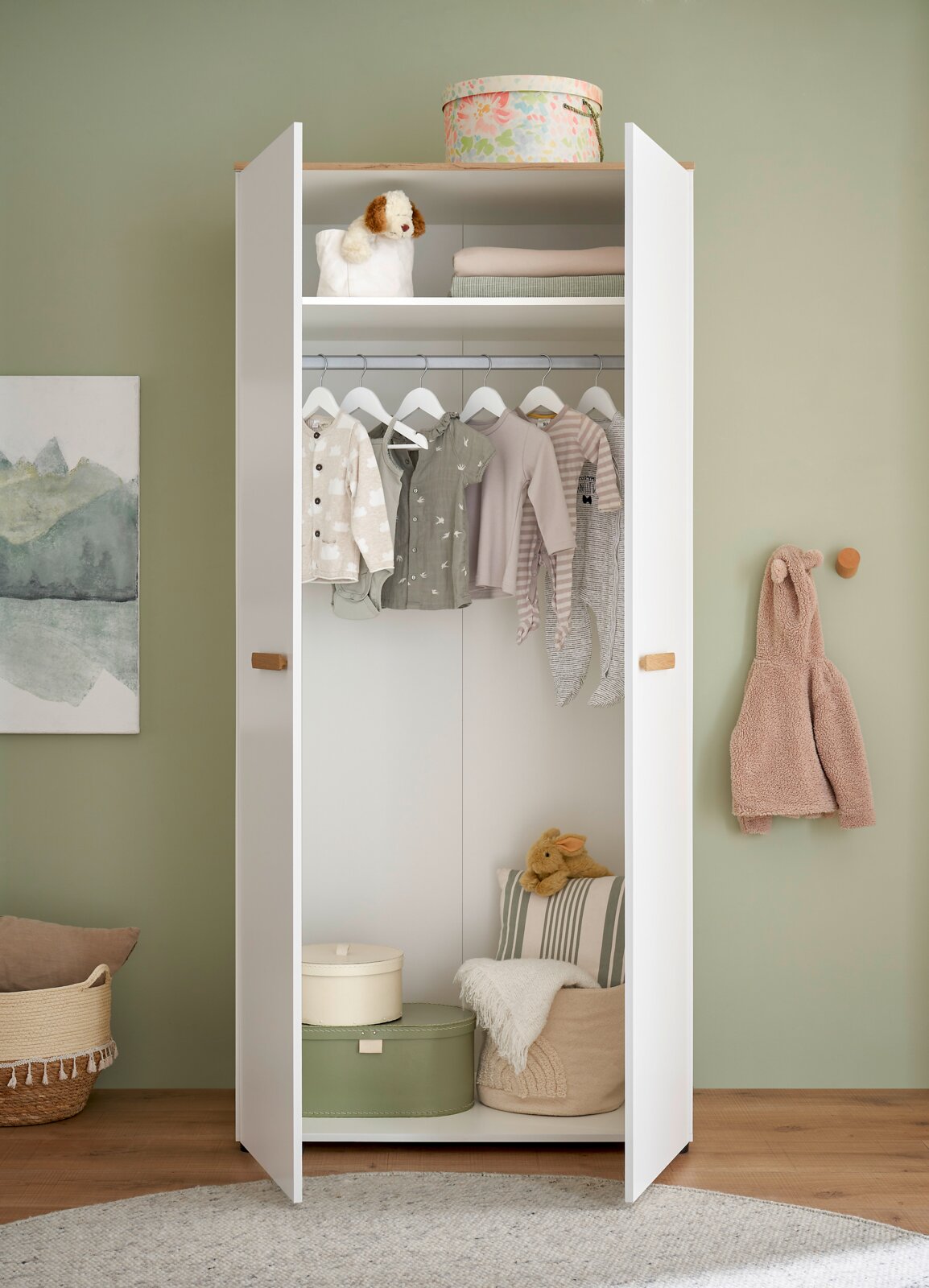 Offener weißer Kinderkleiderschrank mit Holzgriffen, Kleiderstange und Ablagefach, gefüllt mit Babykleidung und Aufbewahrungsboxen; frontale Perspektive