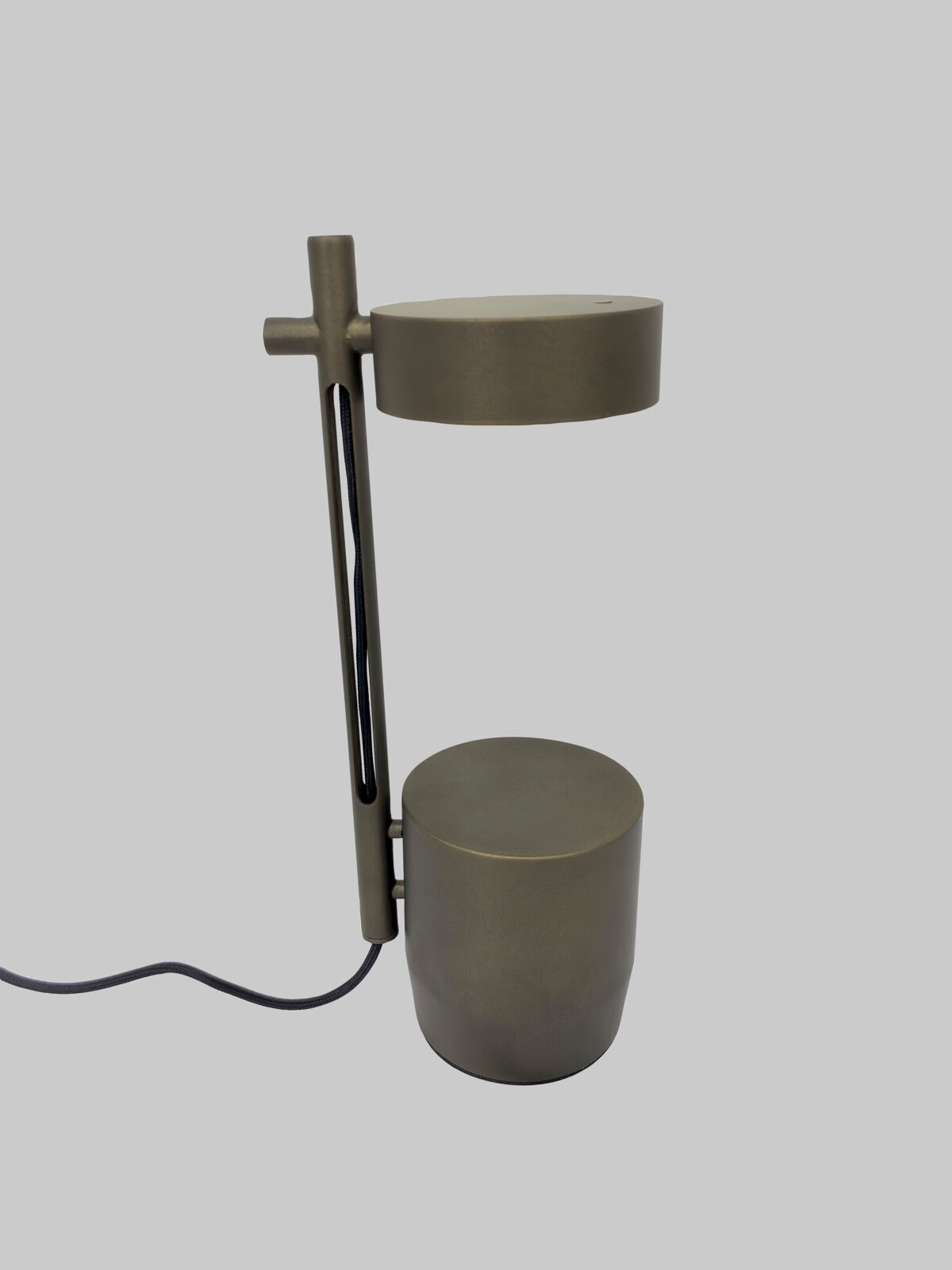 Moderne Tischlampe in Dunkelgrau mit zylindrischem Fuß und rundem Lampenkopf, seitliche Ansicht in leichter Perspektive von vorne, mit sichtbarem Kabel vor hellem Hintergrund
