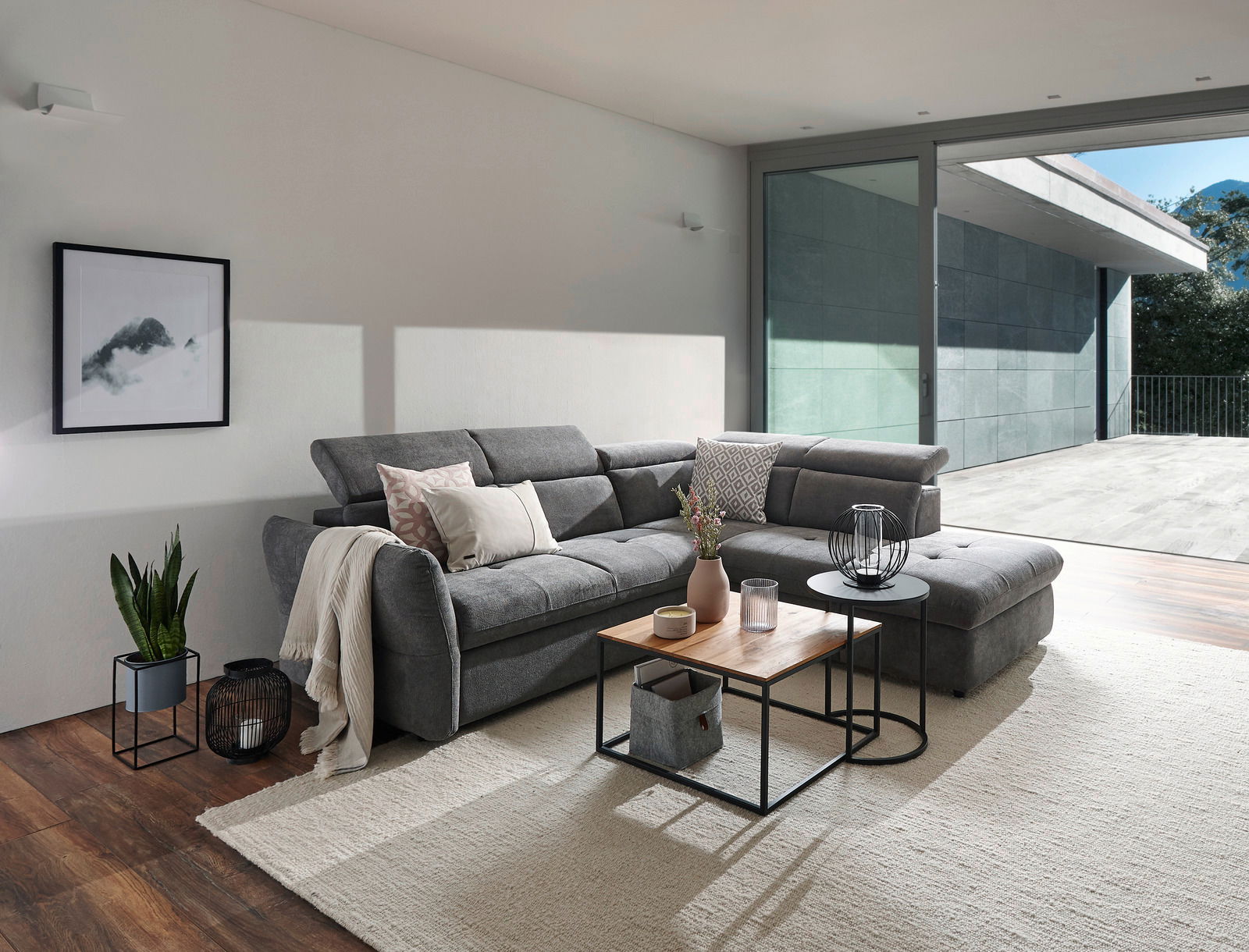 Graues Polsterecksofa MONSERAT in modernem Wohnzimmer, seitliche Perspektive mit Blick auf großen Fensterbereich.