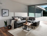 Graues Polsterecksofa MONSERAT in modernem Wohnzimmer, seitliche Perspektive mit Blick auf großen Fensterbereich.