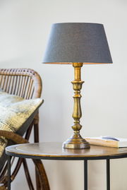 Eleganter Lampenfuß HELGA in Goldoptik mit grauem Lampenschirm, auf einem Tisch neben einem Rattanstuhl, seitliche Perspektive.