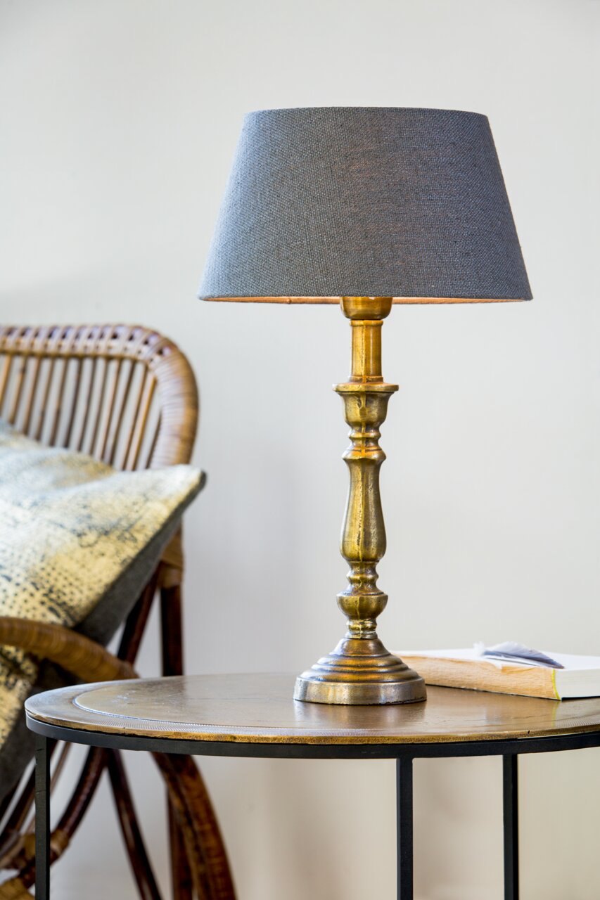 Eleganter Lampenfuß HELGA in Goldoptik mit grauem Lampenschirm, auf einem Tisch neben einem Rattanstuhl, seitliche Perspektive.