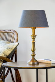 Eleganter Lampenfuß HELGA in Goldoptik mit grauem Lampenschirm, auf einem Tisch neben einem Rattanstuhl, seitliche Perspektive.
