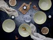 Vier cremefarbene Keramikschalen auf dunkler Tischoberfläche mit goldenem Löffel, Holzbrett, Teelichtern, Stofftüchern und Naturdeko, fotografiert in Draufsicht (Flatlay)