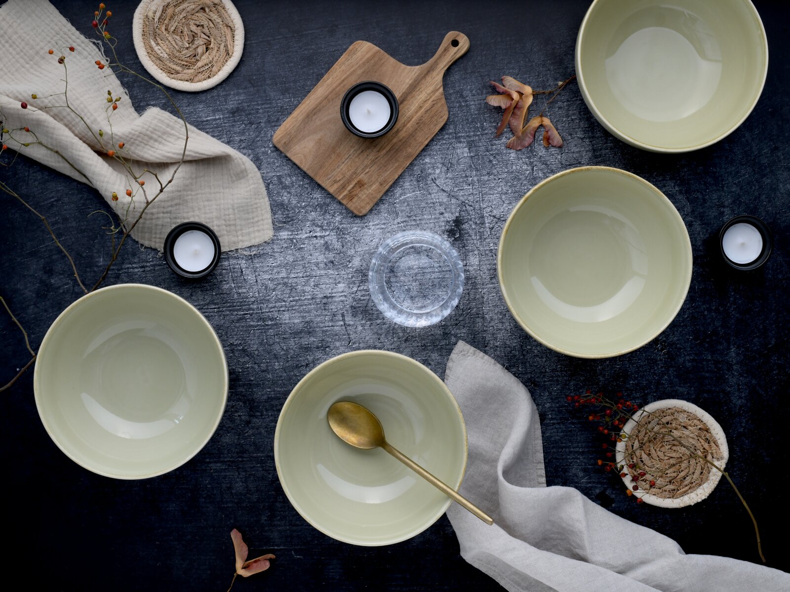 Vier cremefarbene Keramikschalen auf dunkler Tischoberfläche mit goldenem Löffel, Holzbrett, Teelichtern, Stofftüchern und Naturdeko, fotografiert in Draufsicht (Flatlay)