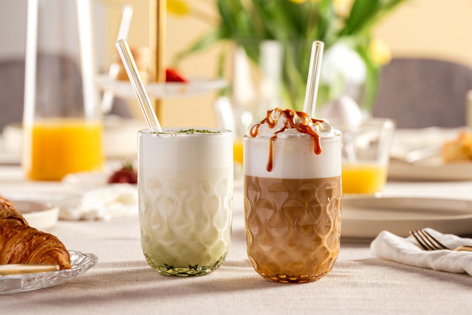 Zwei Gläser mit Matcha Latte und Karamell Latte auf einem gedeckten Tisch, seitliche Perspektive.