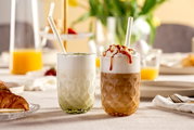 Twee glazen met matcha latte en karamel latte op een gedekte tafel, gezien vanuit de zijkant.