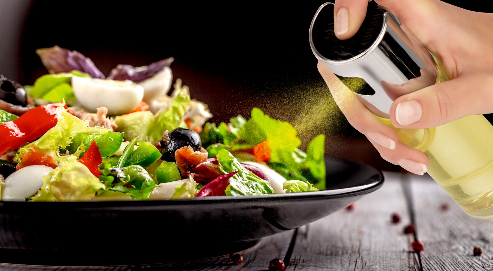 Nahaufnahme aus seitlicher Perspektive: Eine Hand besprüht frischen, bunten Salat mit einem feinen Ölnebel aus einem eleganten Glas-Ölsprüher mit Edelstahlaufsatz.