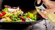 Nahaufnahme aus seitlicher Perspektive: Eine Hand besprüht frischen, bunten Salat mit einem feinen Ölnebel aus einem eleganten Glas-Ölsprüher mit Edelstahlaufsatz.