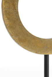 Close-up van een goudkleurig, cirkelvormig ornament op een zwarte standaard, gezien vanaf de zijkant.