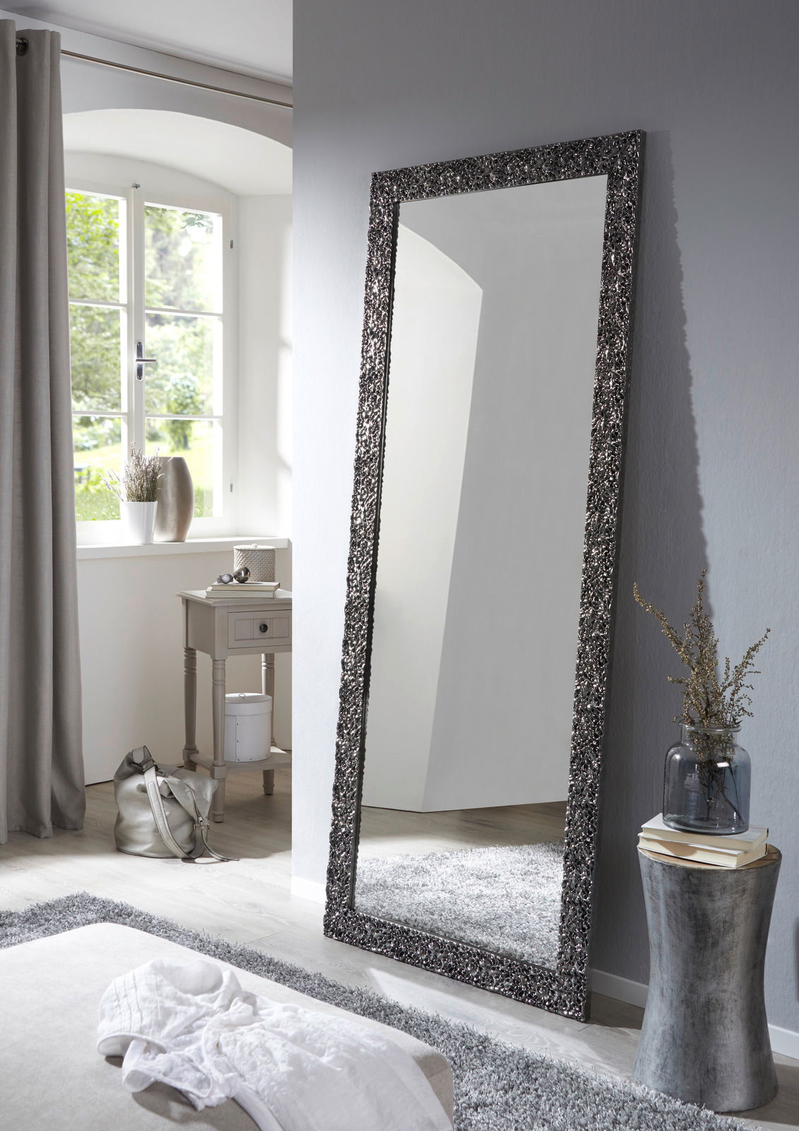 Großer rechteckiger Spiegel mit dekorativem Rahmen, der an einer Wand in einem hellen Raum lehnt, seitliche Perspektive.