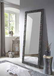 Großer rechteckiger Spiegel mit dekorativem Rahmen, der an einer Wand in einem hellen Raum lehnt, seitliche Perspektive.