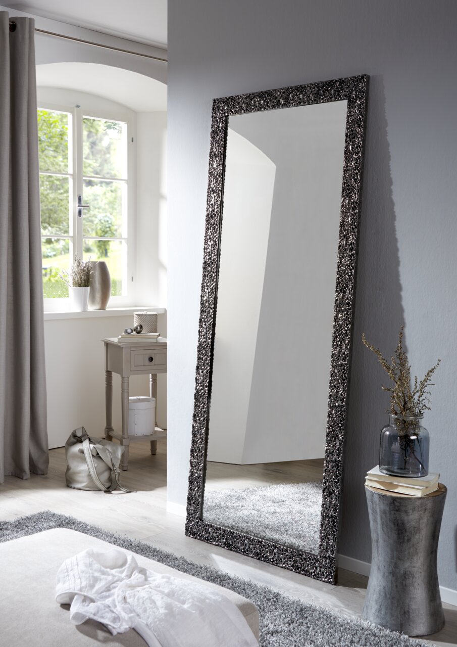 Großer rechteckiger Spiegel mit dekorativem Rahmen, der an einer Wand in einem hellen Raum lehnt, seitliche Perspektive.