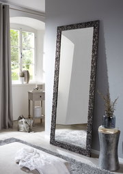 Großer rechteckiger Spiegel mit dekorativem Rahmen, der an einer Wand in einem hellen Raum lehnt, seitliche Perspektive.