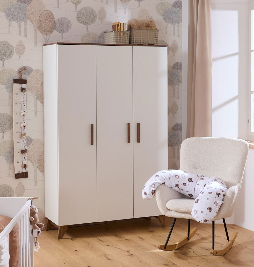 Babyzimmer mit einem dreitürigen weißen Schrank, einem gemütlichen Schaukelstuhl mit Stillkissen und einer Messlatte an der Wand. Perspektive von vorne.