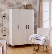 Babyzimmer mit einem dreitürigen weißen Schrank, einem gemütlichen Schaukelstuhl mit Stillkissen und einer Messlatte an der Wand. Perspektive von vorne.