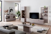 Modernes Wohnzimmer mit taupefarbenem Wohnwand-Set: Lowboard mit Fernseher, Vitrine, Sideboard und Wandregal mit LED-Beleuchtung, aufgenommen aus leicht schräger Frontperspektive auf Augenhöhe.