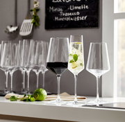 Drei elegante Kelchgläser der Serie Puccini auf einer weißen Oberfläche, gefüllt mit Rotwein, einem Cocktail mit Limette und Minze, und leer, im Vordergrund mit frischen Limetten und Minzblättern, seitliche Perspektive.